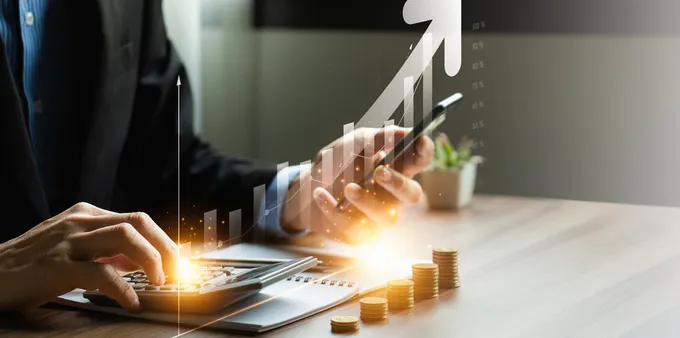concept of rising prices - man calculating expenses while looking at his phone, coins stacked higher and higher in front of him