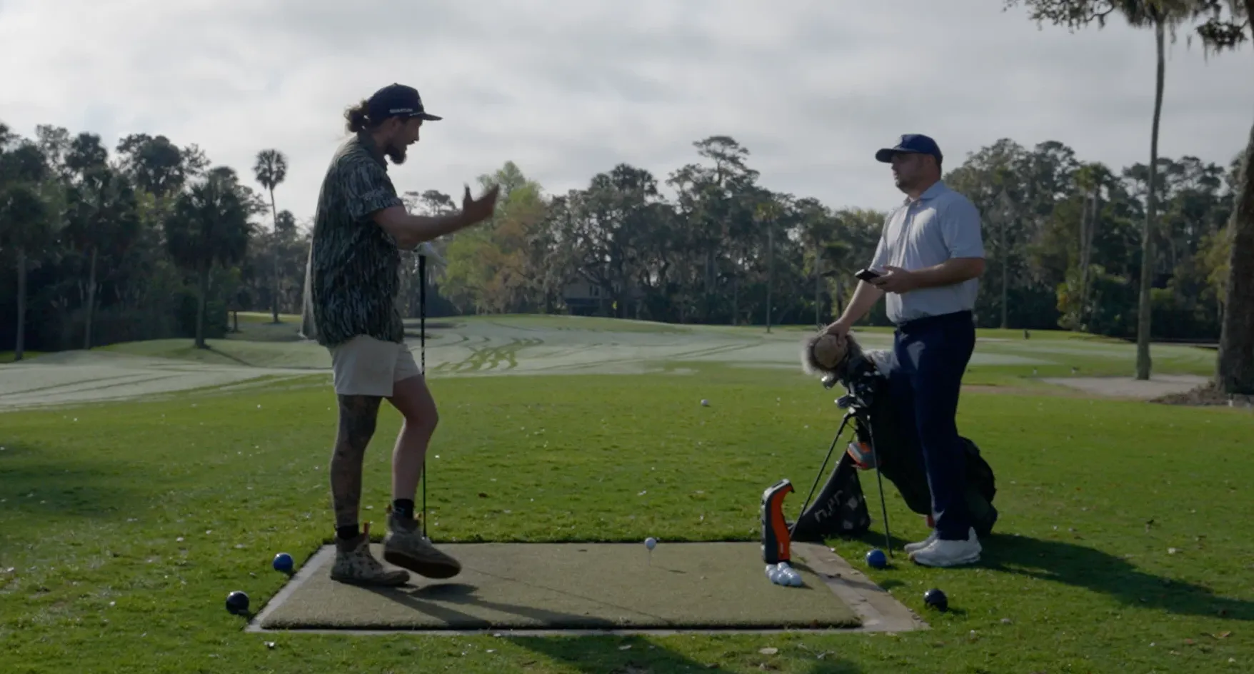 Two men stand on a golf course tee box; one, in shorts and a patterned shirt, gestures animatedly, while the other, in golf attire, stands near a golf bag in a relaxed stance. Trees and fairways are visible in the background.